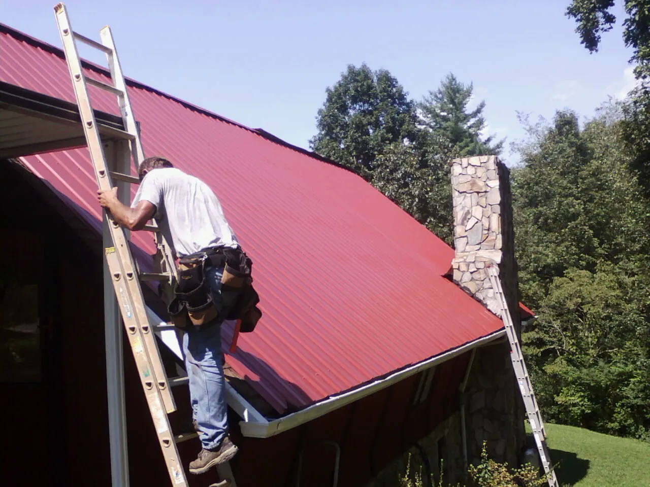 Roofer on a ladder beside a red metal roof and stone chimney — Soffit & Fascia Repair by Roofing Brothers in Lutherville Timonium